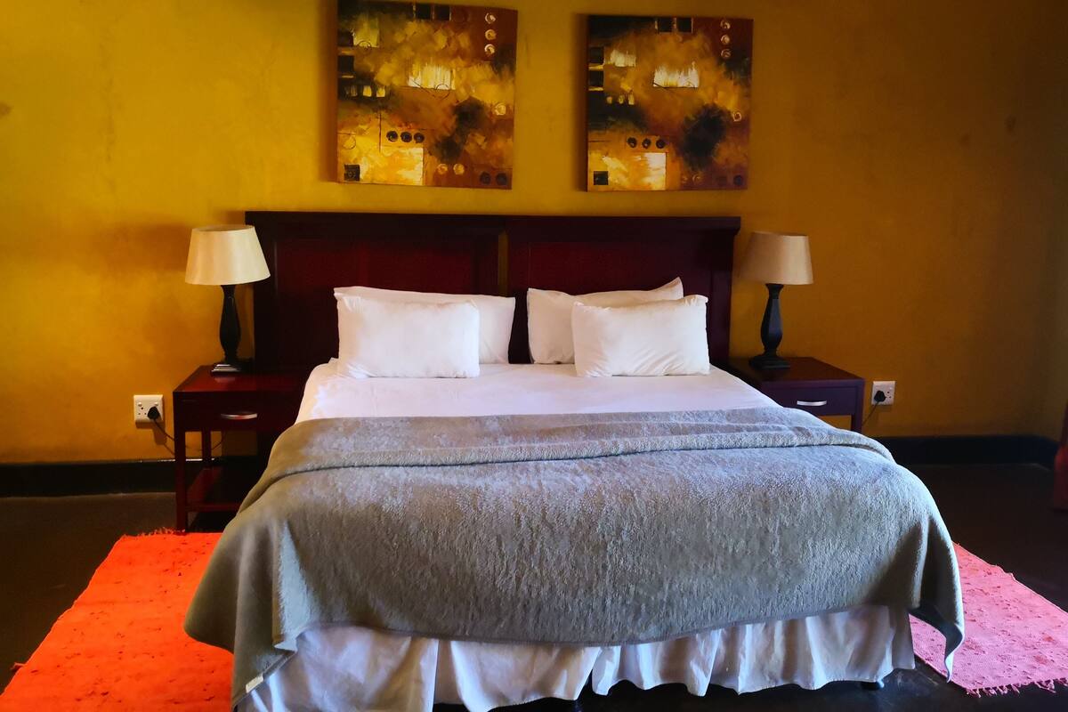 A neatly made bed with white linens and a grey blanket, flanked by two bedside tables with lamps, in a room with yellow walls and two abstract paintings above the bed.