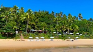 Una playa con sillas y sombrillas blancas, rodeada de palmeras y cabañas de playa.