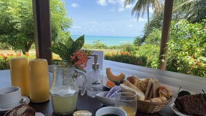 Una mesa servida con alimentos para el desayuno, incluyendo pasteles, pan y un bol de fruta, con vista al océano y follaje al fondo.
