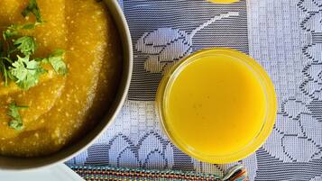 En skål suppe pyntet med koriander, to glas med en gul væske og en hvid tallerken på en farverig vævet måtte.