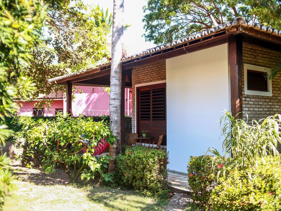 A small brick house with a tiled roof, surrounded by lush greenery and a palm tree.