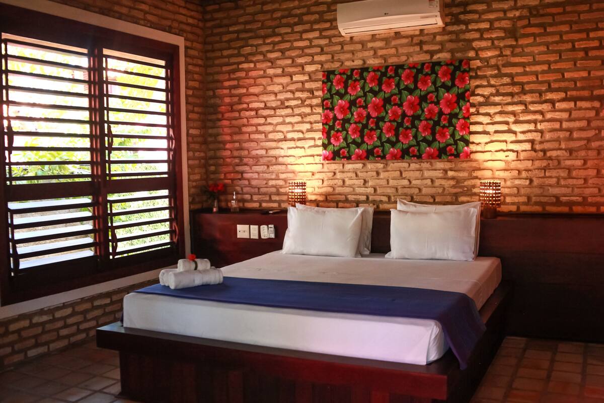 Een kamer met een bakstenen muur, een bed met wit beddengoed, een raam met zonwering en een aan de muur bevestigde airconditioning.