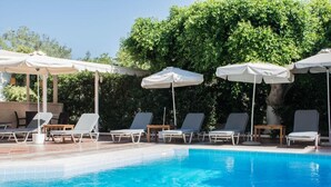 A swimming pool with lounge chairs and umbrellas, surrounded by greenery. - Rena Apartments Gouves (Hersonissos)