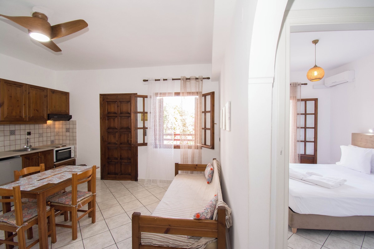 Una habitación compacta con cama, una pequeña mesa con sillas, kitchenette y ventilador de techo.