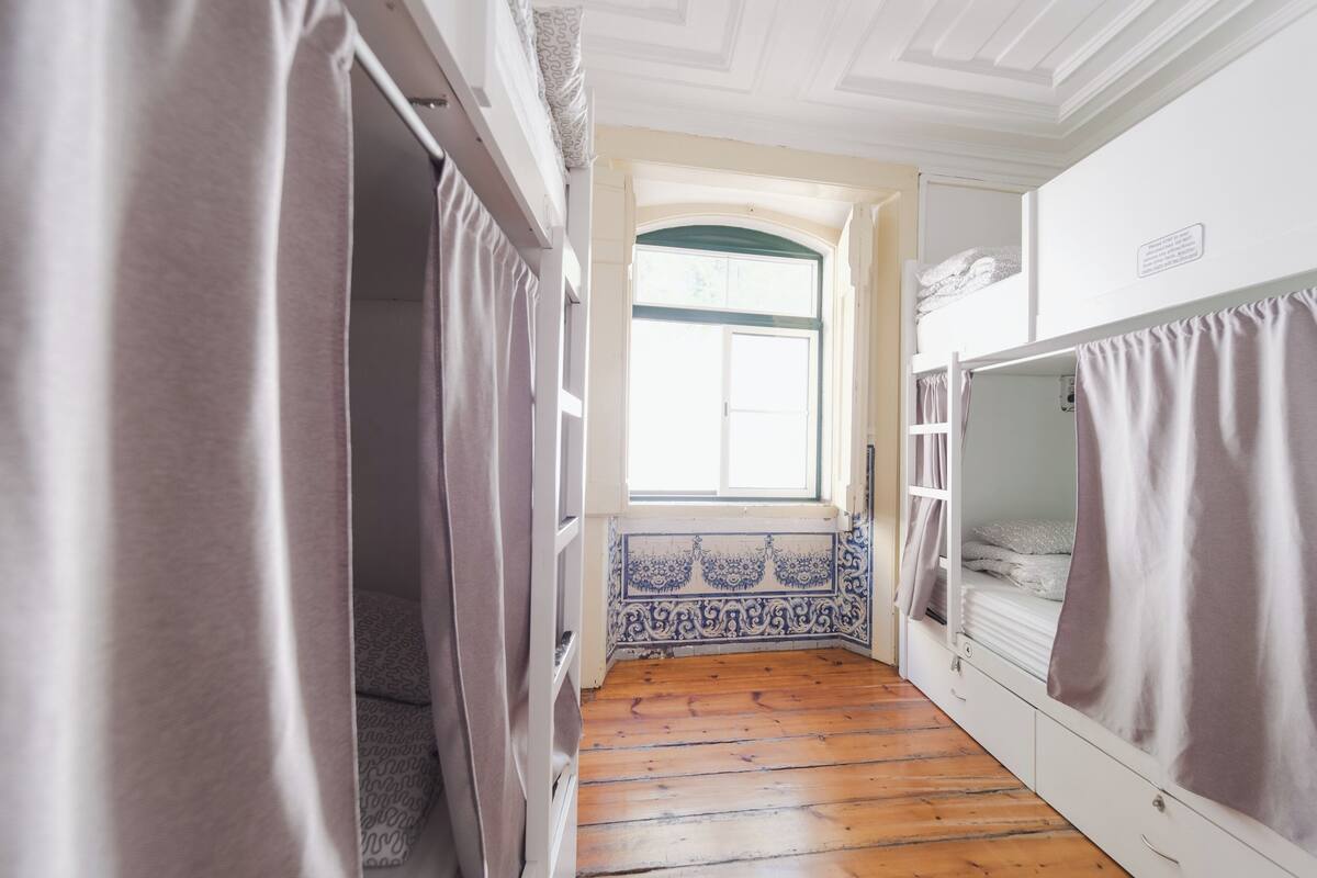 Una habitación estrecha con suelo de madera, una ventana con marco verde y una cama blanca con colcha gris.