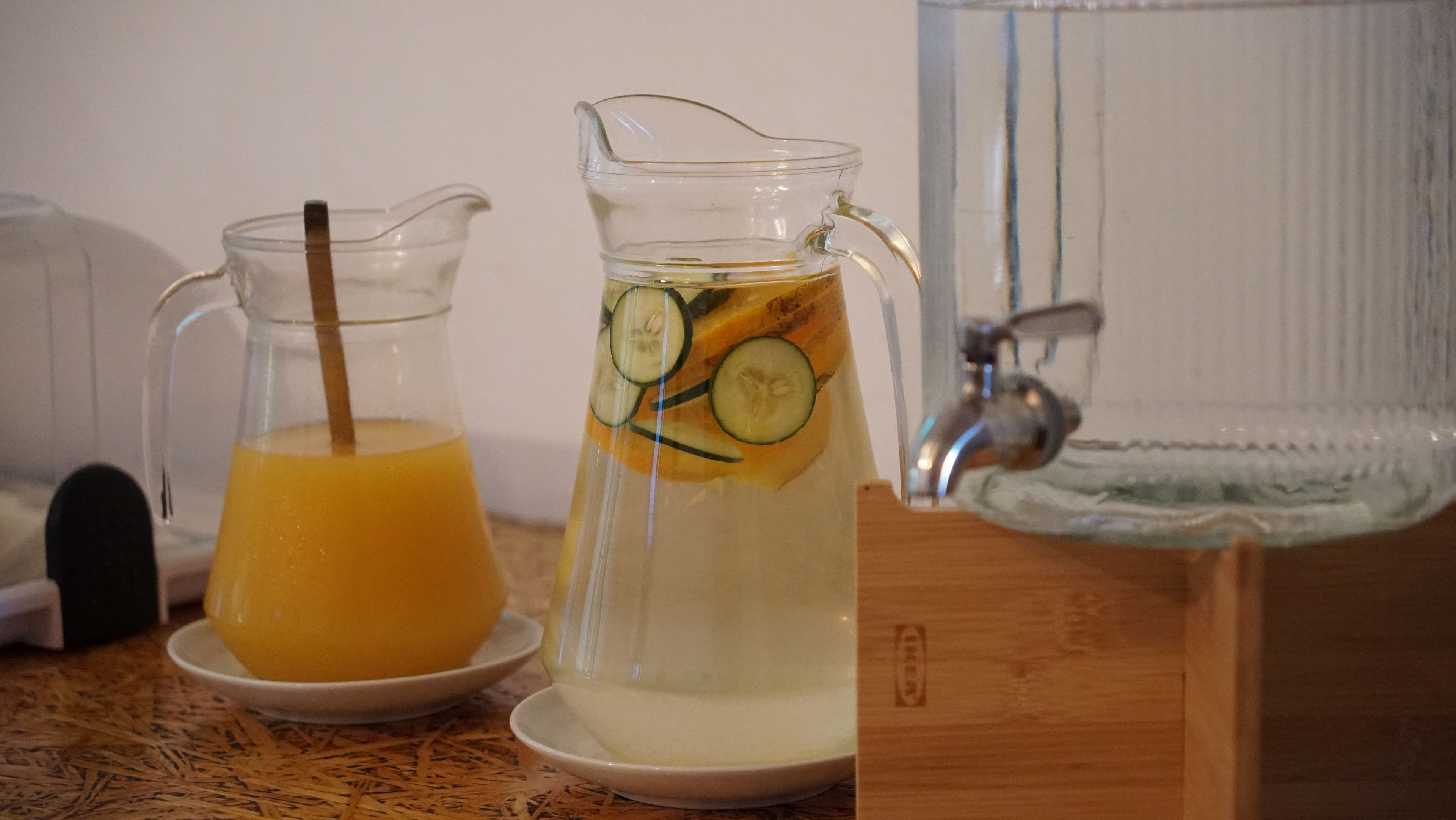 A pitcher with a straw and sliced cucumbers, a glass pitcher with a straw, and a water dispenser on a wooden surface.