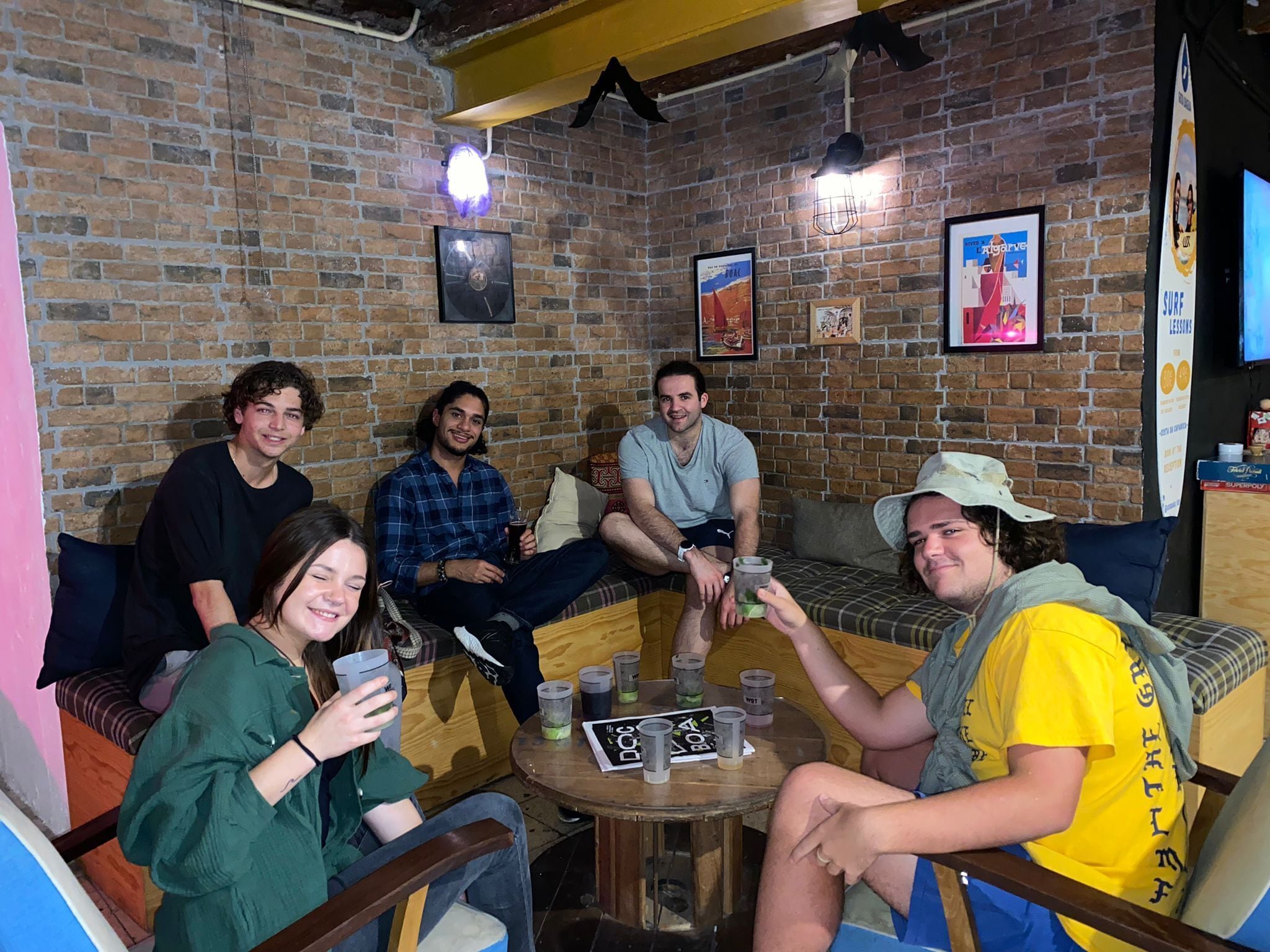 Un groupe de personnes est assis autour d’une table en bois, dans une pièce aux murs de briques, meublée de meubles en bois et ornée de divers tableaux encadrés.