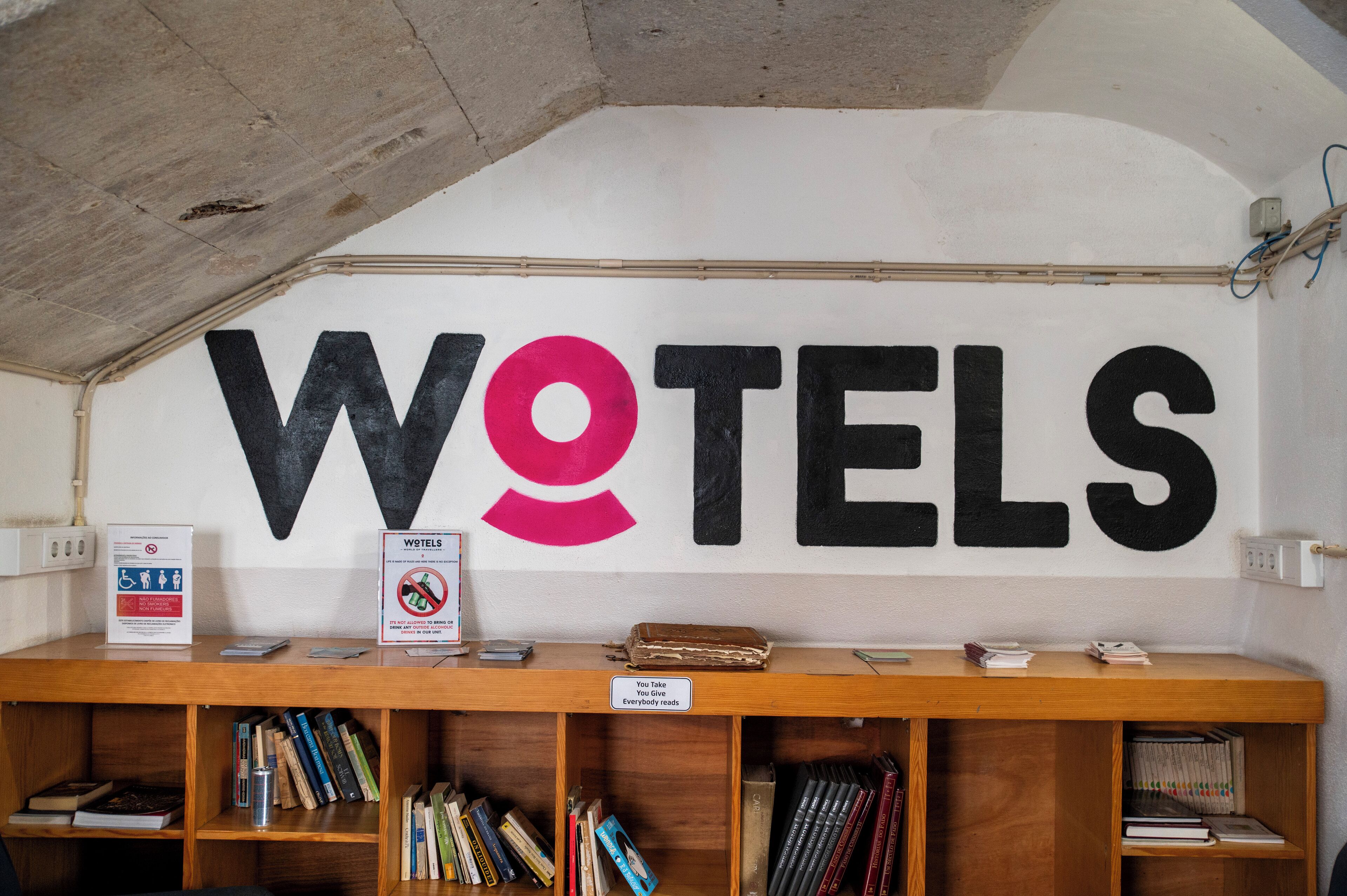 Una pared con la palabra "WOTELS" en letras grandes, un cartel que prohíbe fumar y un letrero que dice "Toma, devuelve."