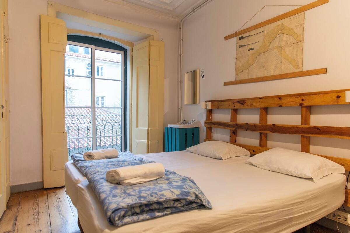 Un dormitorio con cabecera de madera, cama con ropa de cama blanca, manta azul y blanca estampada, un pequeño armario azul y un mapa en la pared.