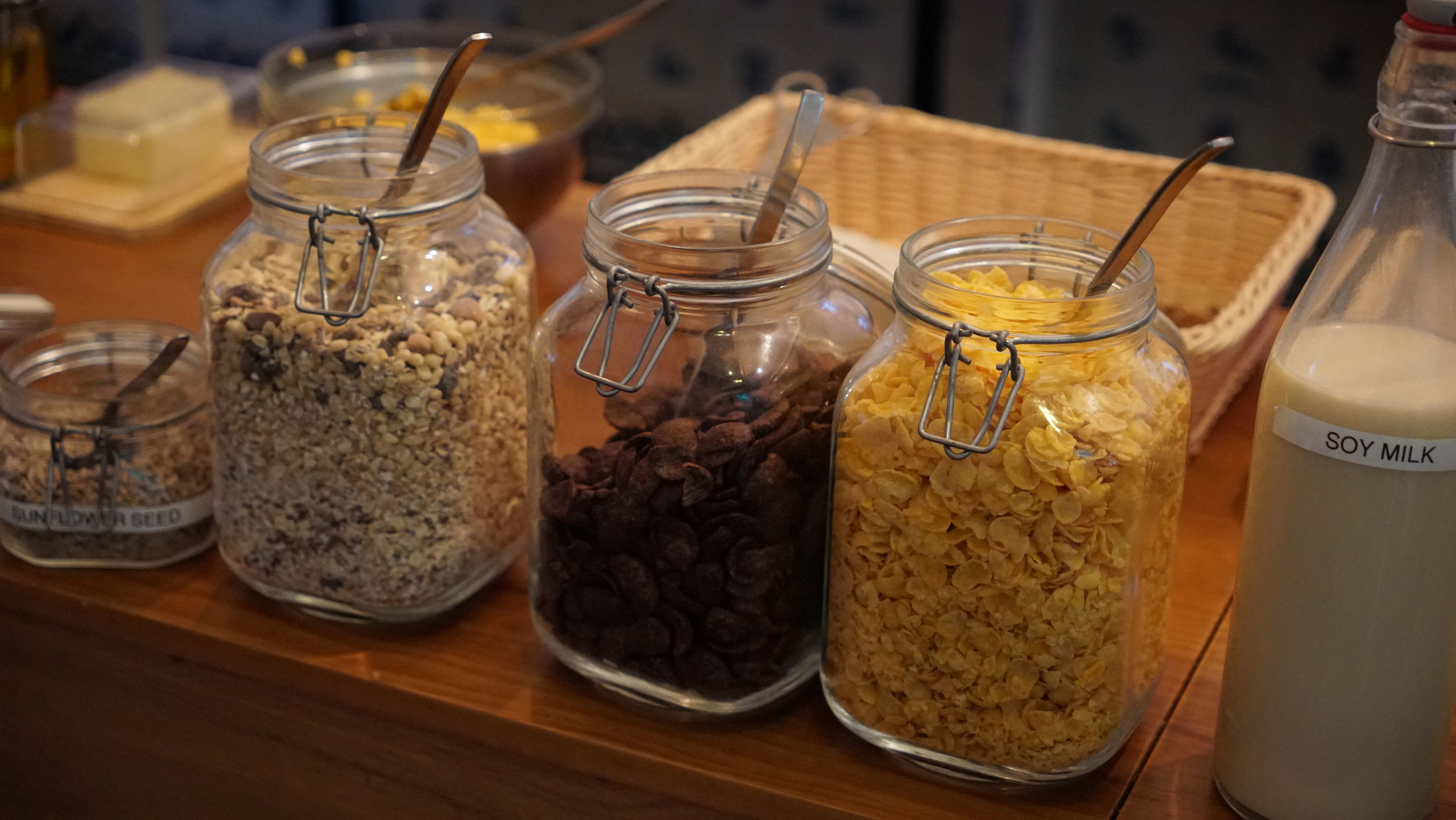 Une variété d’aliments en conserve dans des bocaux en verre, notamment des céréales et du lait de soja, sont disposés sur une table en bois.