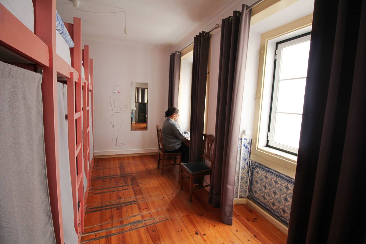 Una habitación con piso de madera, un escritorio y una silla. Hay una ventana con cortinas, un espejo y una toalla colgada en un perchero.
