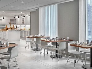 Un comedor moderno con sillas y mesas blancas, una cocina al fondo y grandes ventanas con cortinas ligeras.