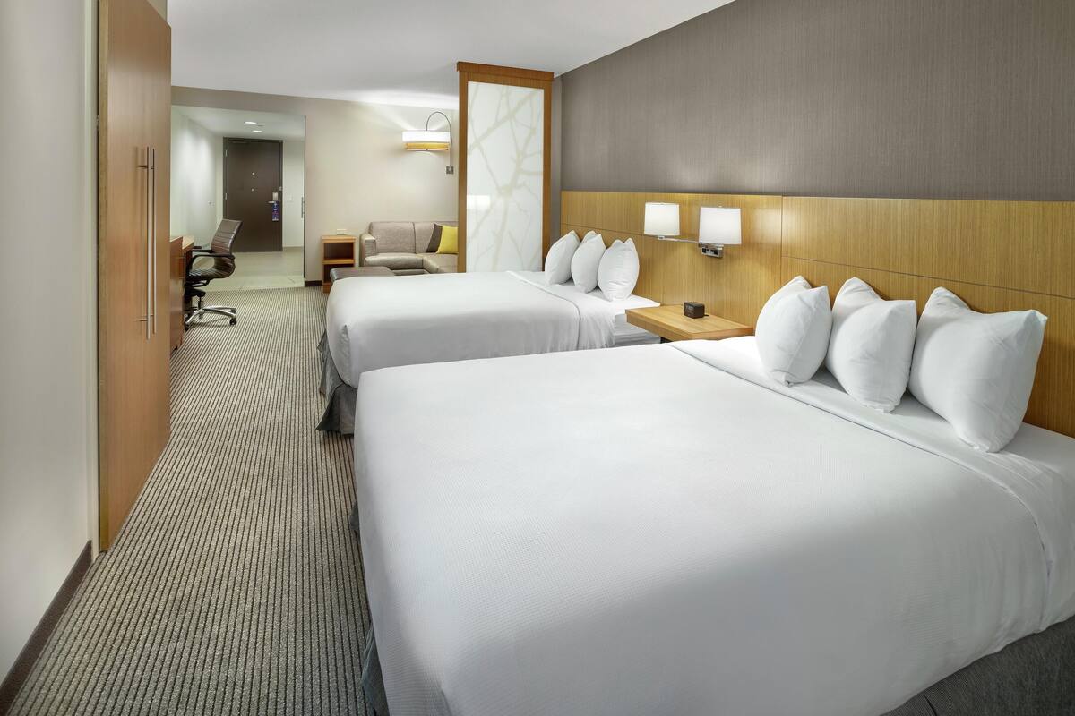 A hotel room with two beds, a desk, a chair, and a small table with a lamp.