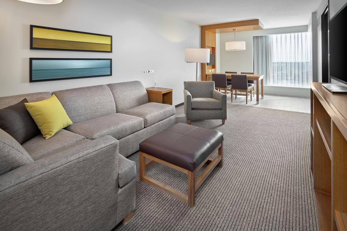 A modern hotel room with a grey sofa, a brown ottoman, a dining area with a table and chairs, a flat-screen TV, and a large window with curtains.