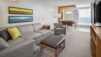 Una habitación de hotel moderna con un sofá gris, un reposapiés marrón, un área de comedor con mesa y sillas, un televisor de pantalla plana y un amplio ventanal con cortinas.