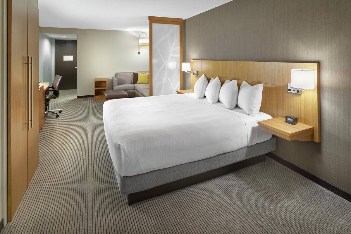 A hotel room with a large bed, a desk with a lamp, a chair, and a small table.