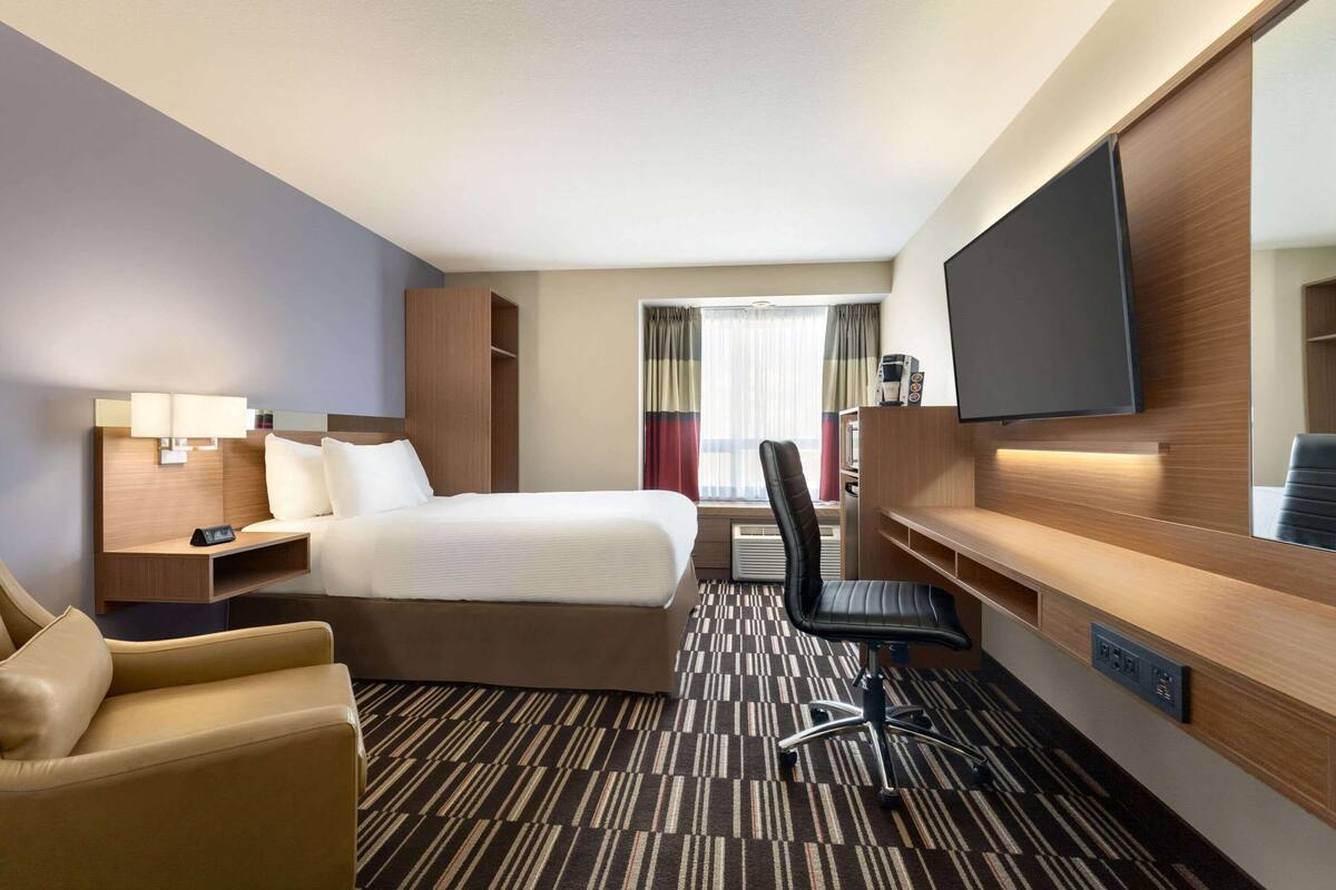 Una habitación de hotel con cama, escritorio, silla, televisión y ventana con cortinas.