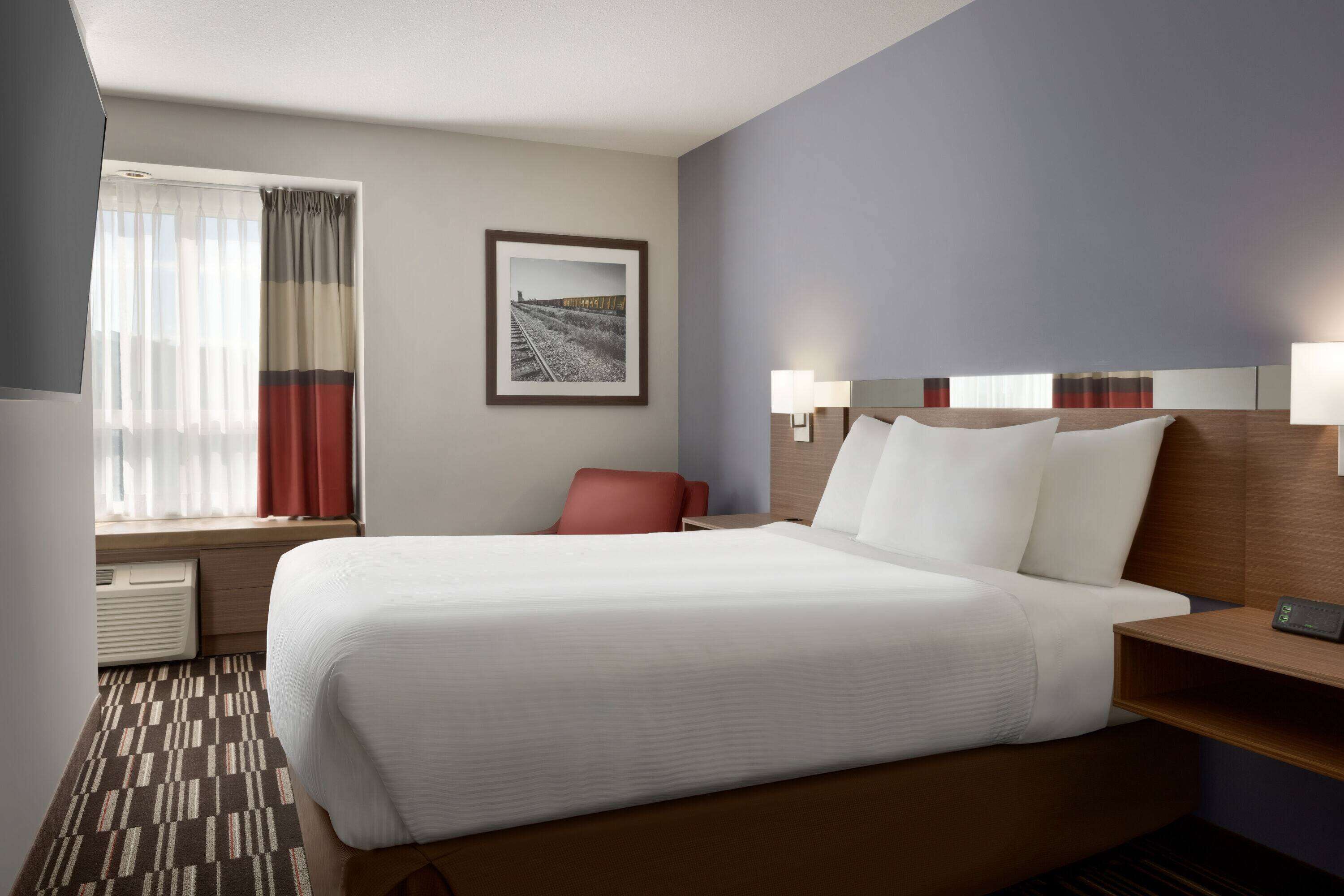 A hotel room with a large bed, a desk with a lamp, a chair, a window with curtains, and a framed picture on the wall.