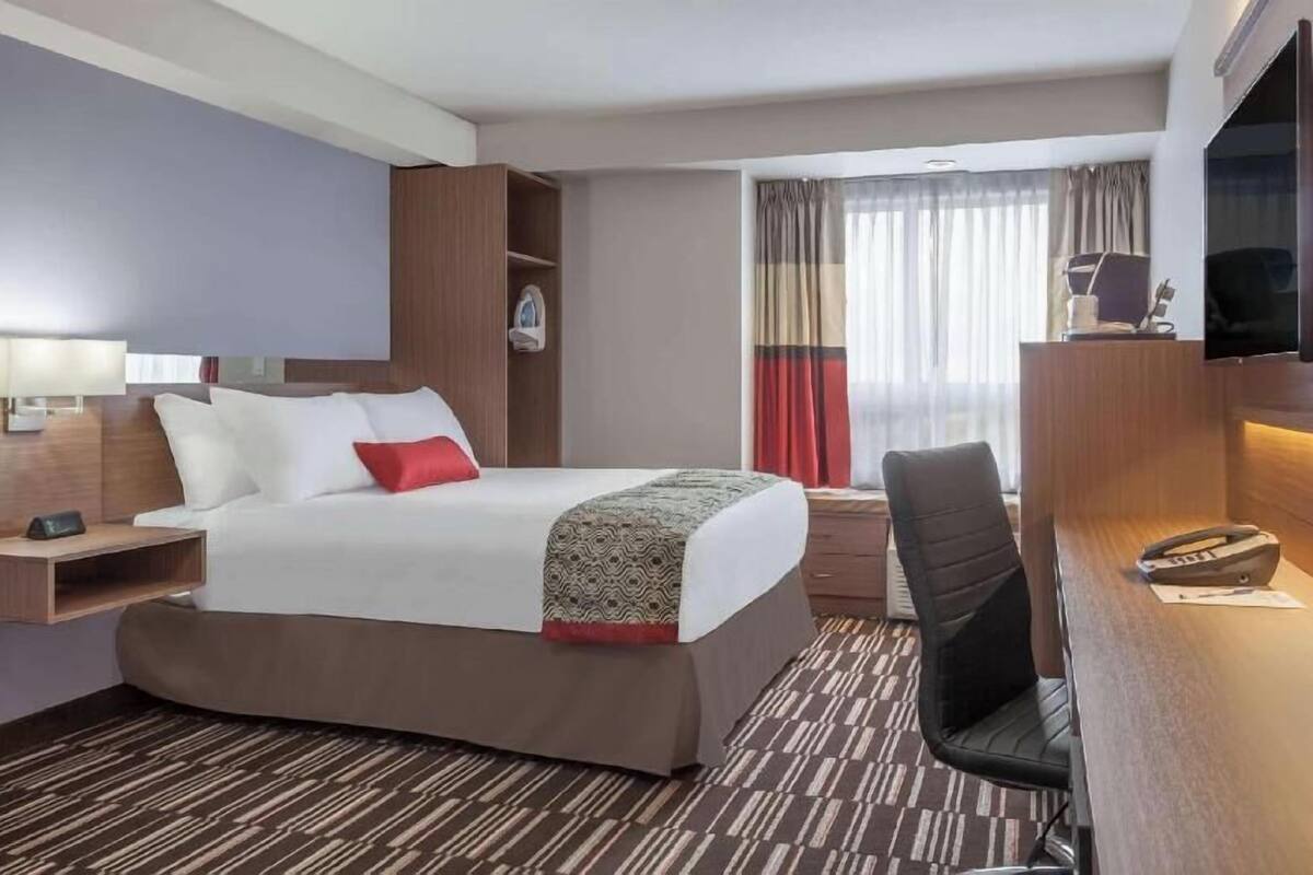 Habitación de hotel con cama, escritorio, silla, televisor y ventana con cortinas.