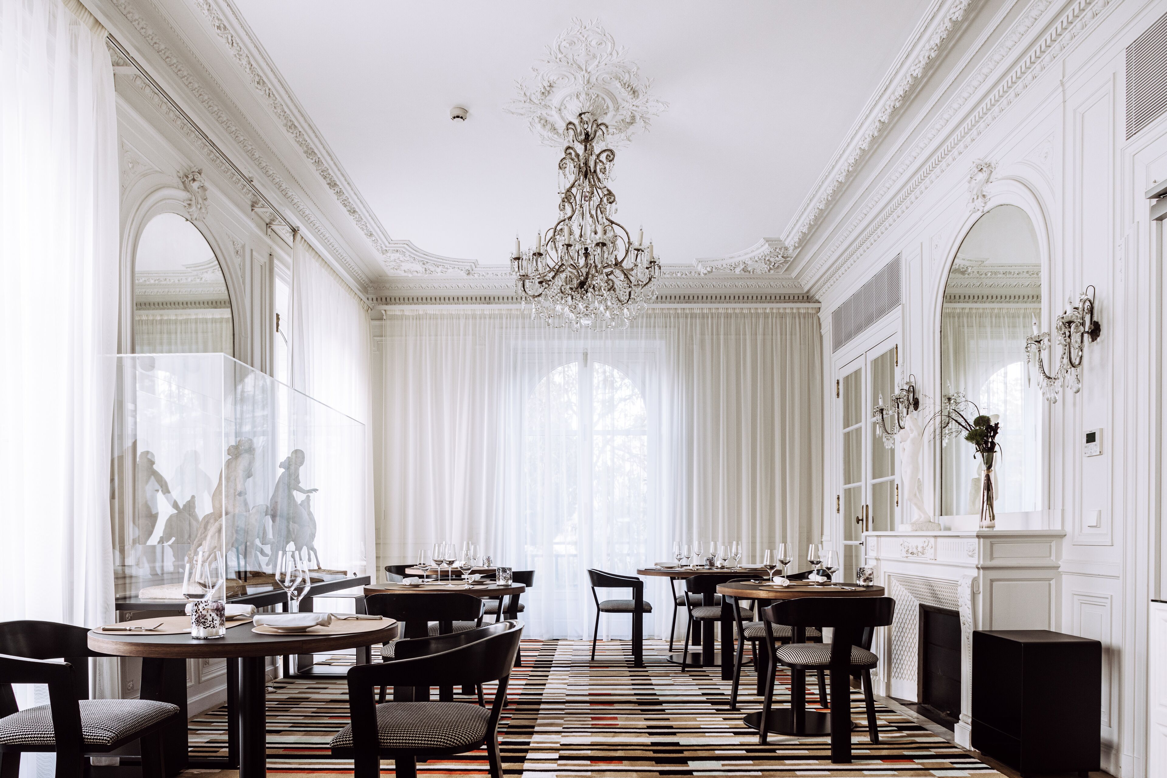 A formal dining room with ornate chandeliers, large windows with sheer curtains, and a patterned floor.