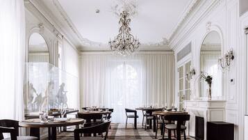 A formal dining room with ornate chandeliers, large windows with sheer curtains, and a patterned floor.