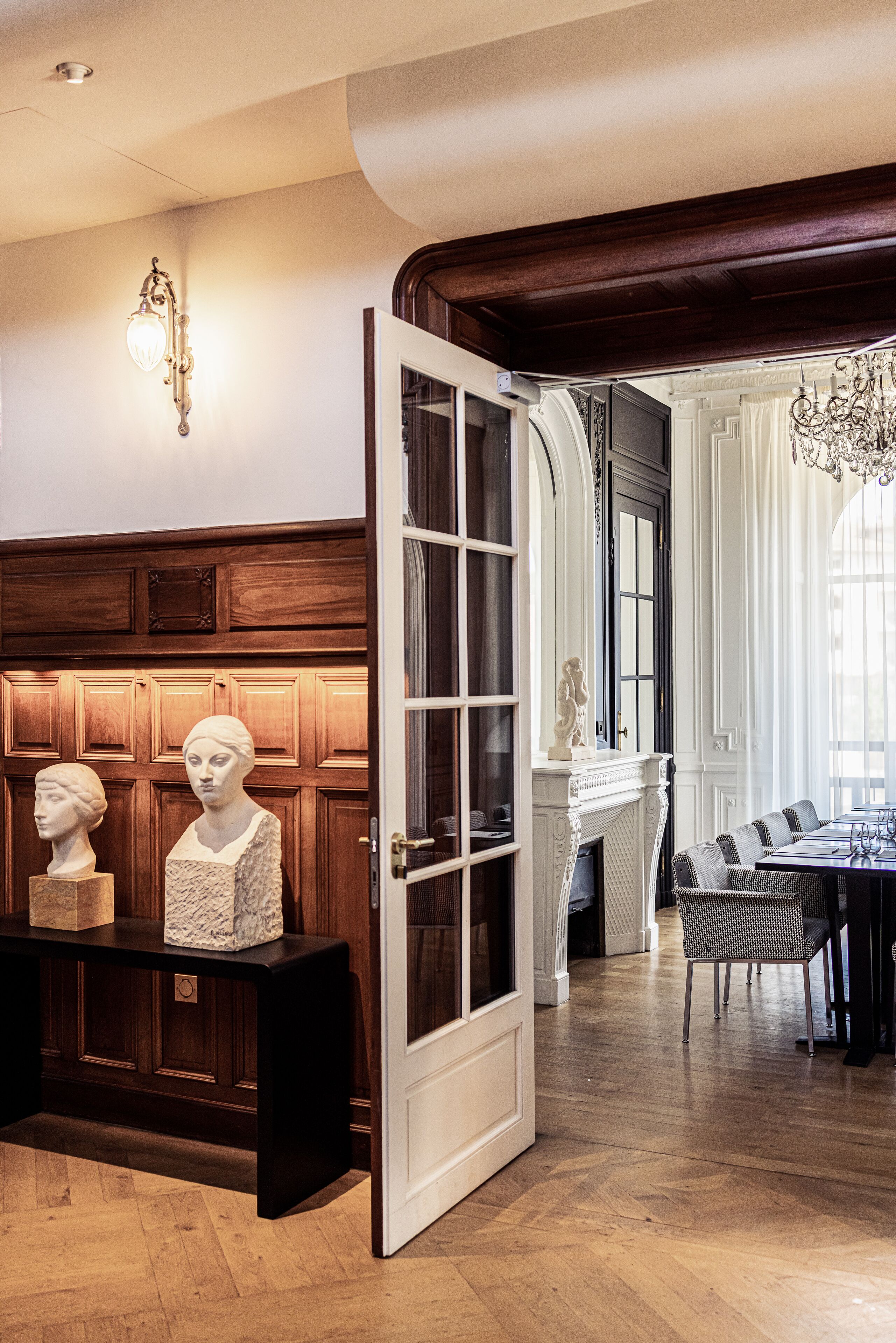 Un hall majestuoso con un revestimiento de paneles de madera, una puerta blanca y un banco adornado con bustos. Se observa un candelabro y un comedor con una mesa y sillas al fondo.