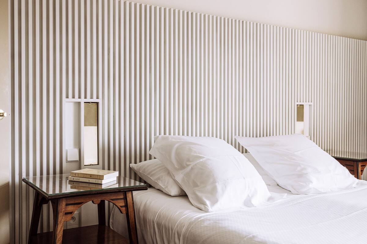 Una habitación de hotel con papel tapiz a rayas, una cama con sábanas blancas, una mesita de noche con un libro y una lámpara.