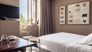 A hotel room with a bed, a desk with a kettle, a chair, a TV, and framed artwork on the wall.