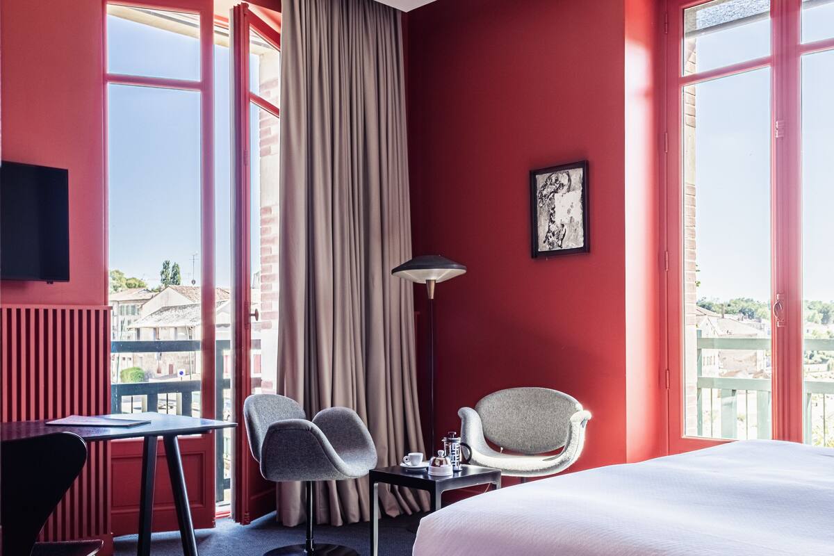 Habitación de hotel con una cama grande, dos sillas, una mesita con lámpara y vistas a edificios a través de las ventanas.