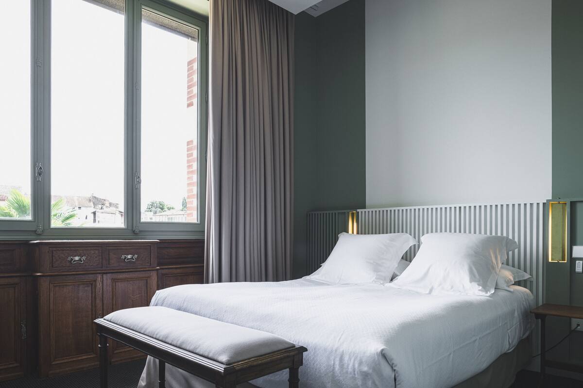 Una cama bien hecha con ropa blanca, un banco de madera y una mesita de noche con una lámpara.