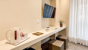 A modern hotel room with a white console table, a flat-screen TV mounted on the wall, a kettle, a small plant, and a window with sheer curtains. - Ecce Roma Suites (Rome)