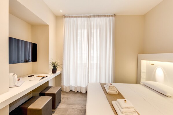 A modern hotel room with a large bed, a bedside table with a lamp, a TV mounted on the wall, and a window with white curtains. - Ecce Roma Suites (Rome)