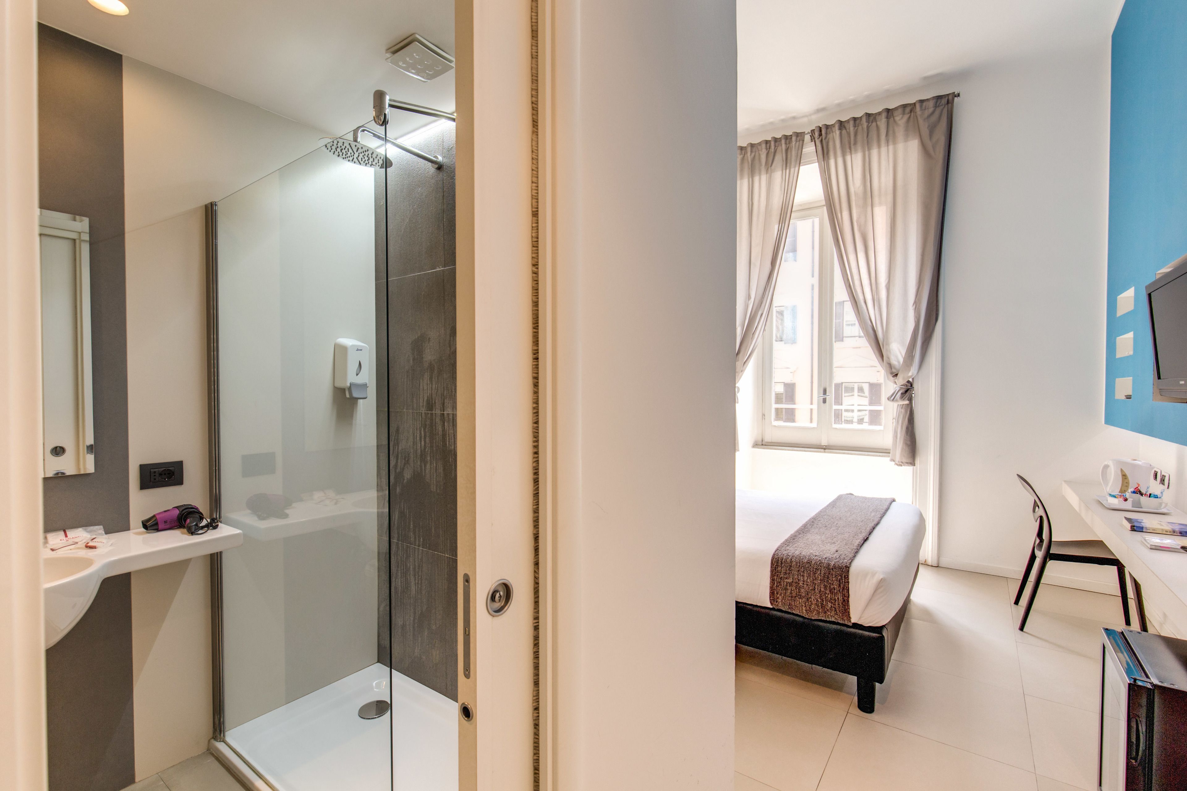 Camera d'albergo con cabina doccia in vetro, un letto con coperta marrone, una scrivania con sedia e una finestra con tende.