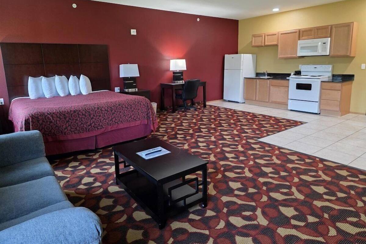 A hotel room with a bed, a desk, a chair, a small table, a microwave, a refrigerator, and a kitchenette.