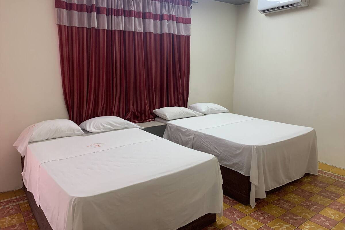 Una habitación con dos camas individuales, un ventanal con cortinas rojas, un equipo de aire acondicionado en la pared y piso de baldosas.