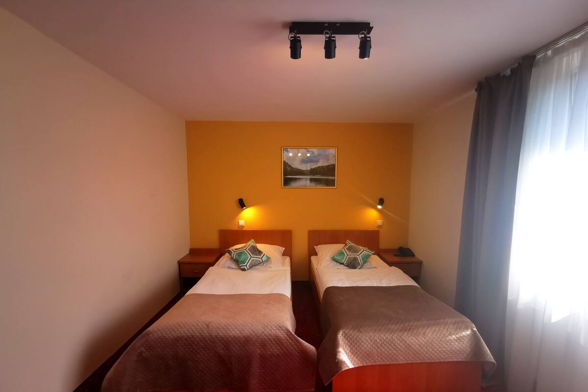 A hotel room with two single beds, a nightstand with a lamp, a painting on the wall, and a window with curtains.