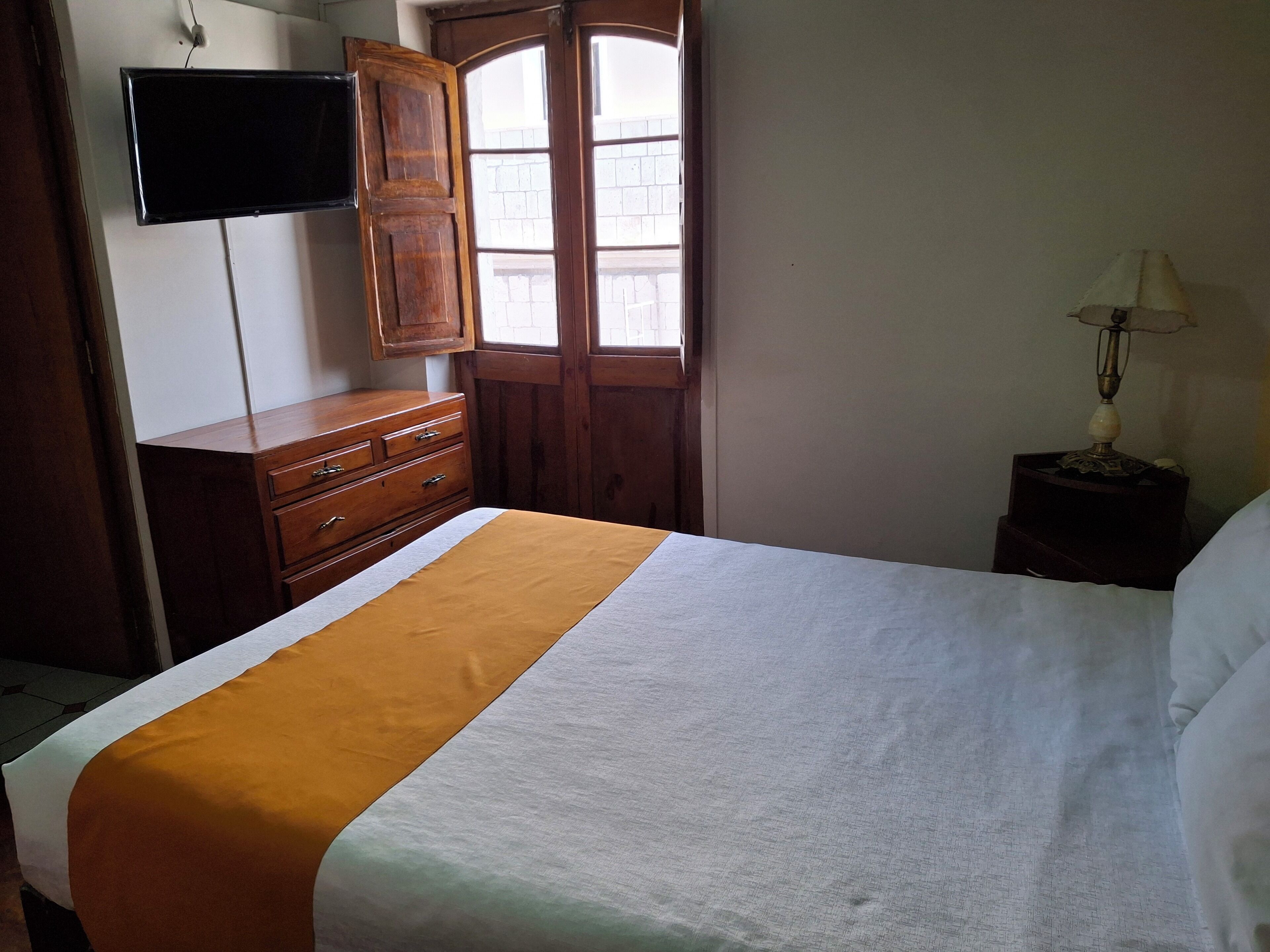 A bedroom with a bed, a wooden dresser, a television mounted on the wall, and a window with a view of buildings outside.