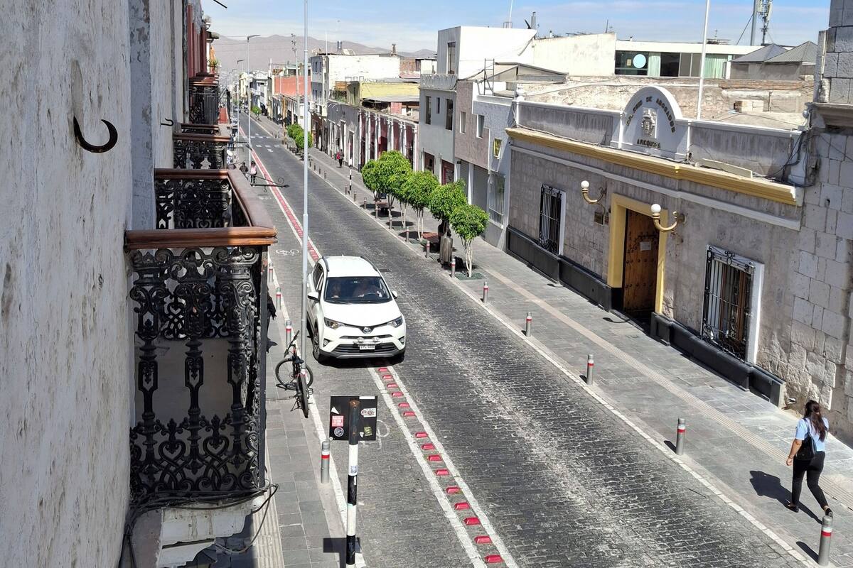 Vista d'un carrer amb un cotxe blanc que avança per un camí de pedra, amb edificis als costats que tenen balcons i un cel blau clar.