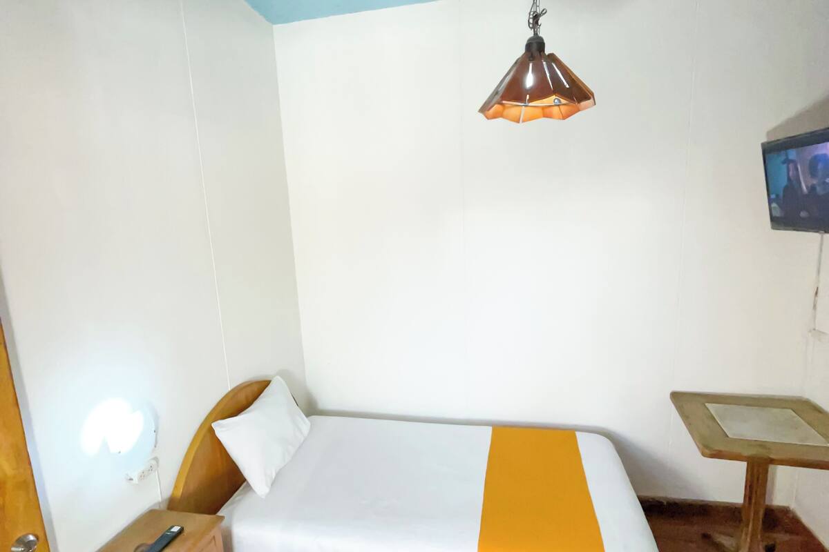 Habitació petita i senzilla amb un llit individual, una taula de nit, una petita taula i un televisor fixat a la paret.