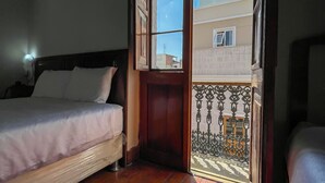 Un dormitorio con piso de madera, una cama con ropa de cama blanca y vista a un edificio a través de un puerta abierta.