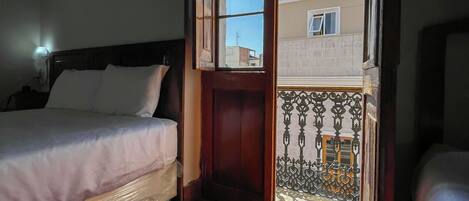 Un dormitorio con piso de madera, una cama con ropa de cama blanca y vista a un edificio a través de un puerta abierta.