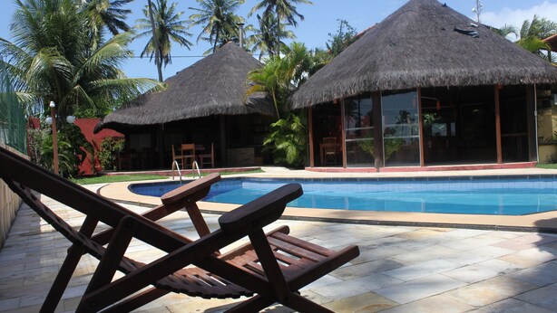 A pool area with thatched-roof structures, lounge chairs, and palm trees. - Pousada Corais do Sul (Tibau do Sul)