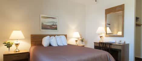 A hotel room with a bed, two bedside tables with lamps, a desk with a chair, a mirror, and a painting on the wall.