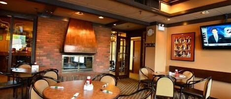 A restaurant with round tables, chairs, a brick fireplace, and a TV mounted on the wall.