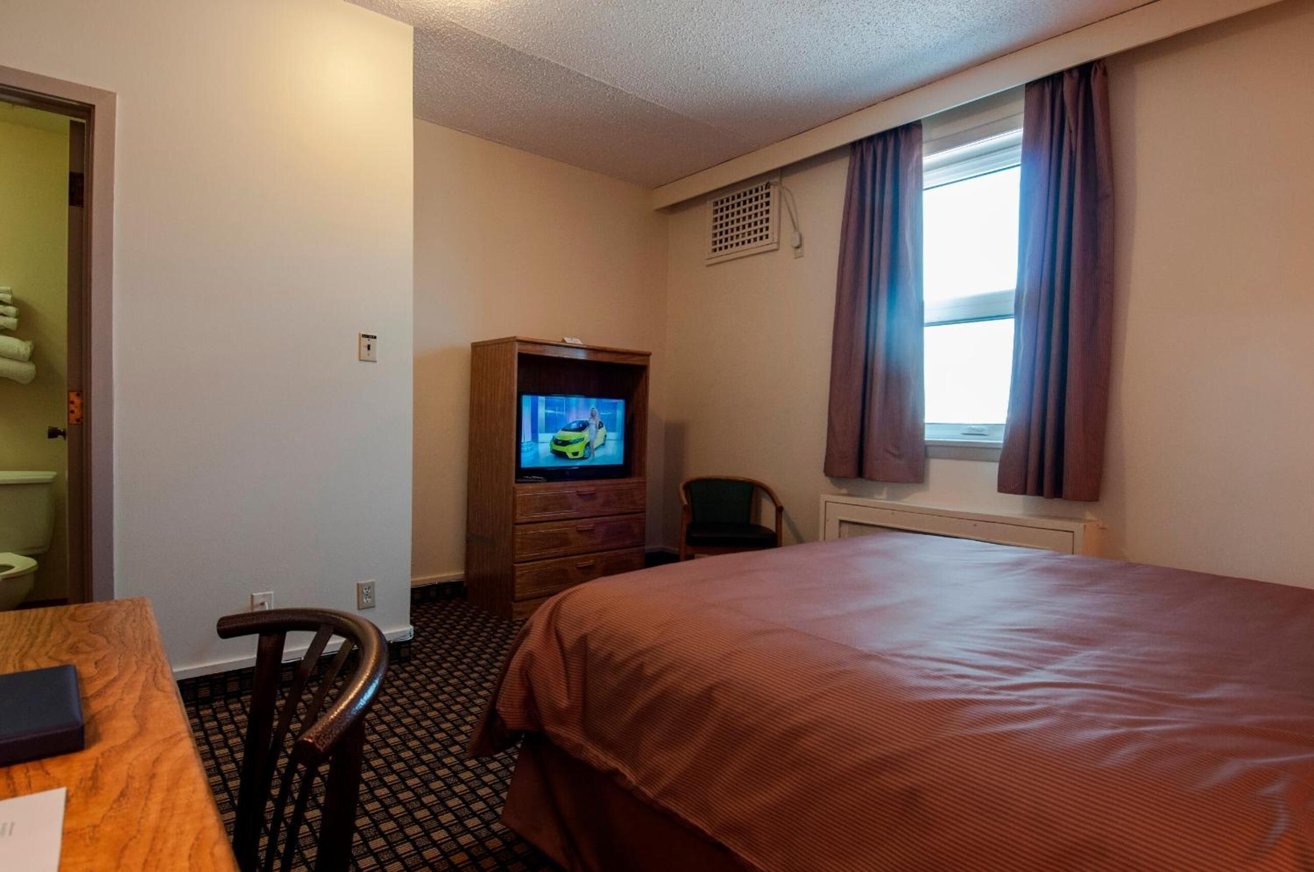 Une chambre d’hôtel avec un lit, un téléviseur posé sur une commode en bois, une chaise et une fenêtre avec des rideaux.