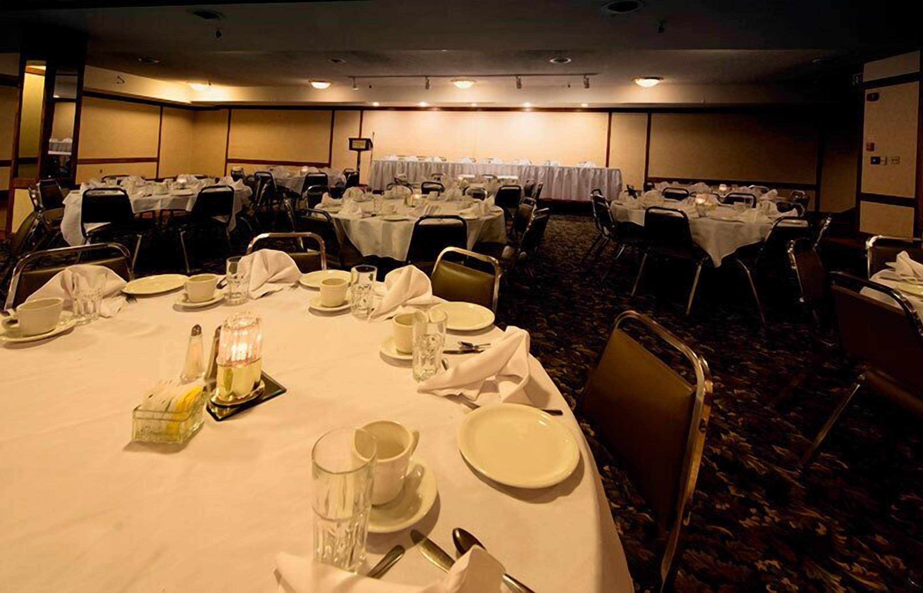A banquet hall with multiple round tables set for dining, each with white tablecloths, plates, glasses, and cutlery.
