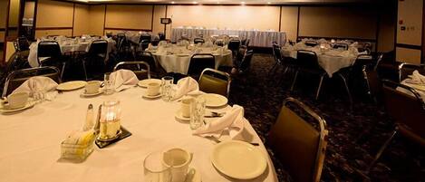 A banquet hall with multiple round tables set for dining, each with white tablecloths, plates, glasses, and cutlery.