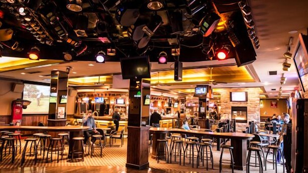 A bar area with multiple stools and tables, a counter with a bartender, and overhead lighting fixtures. - Curtis Gordon Motor Hotel (Winnipeg)