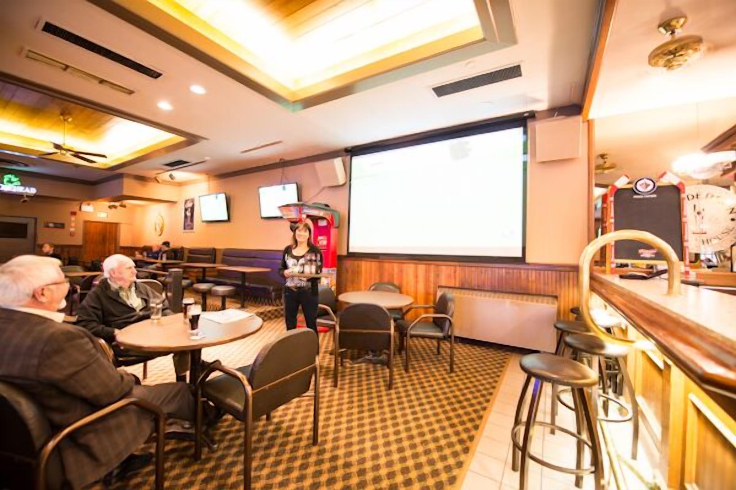 Un restaurante con mesas redondas, taburetes de bar, una pantalla de proyección y un ventilador de techo.