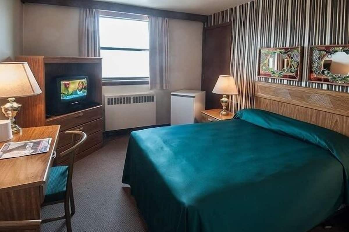 A hotel room with a bed, a desk with a chair, a television, a radiator, and a window with curtains.